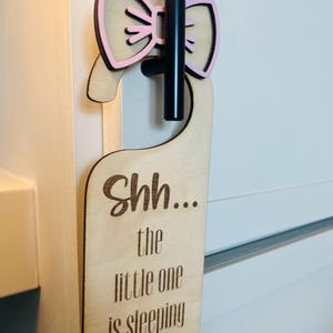 May include: Wooden door hanger with a pink bow accent. The text "Shh... the little one is sleeping" is engraved on the light-colored wood. The hanger is designed to be placed on a door handle.