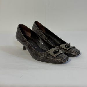 Peut inclure: Une paire d'escarpins gris foncé avec un motif texturé en peau de serpent. Ces chaussures présentent un petit talon, un bout carré et une boucle rectangulaire métallique proéminente sur le devant. La doublure intérieure est noire avec du texte.