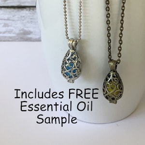 May include: Two essential oil diffuser necklaces with ornate, openwork pendants hang from a white mug. One pendant is silver with a blue interior, the other is bronze with a yellow interior. The text "Includes FREE Essential Oil Sample" is visible.