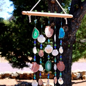 Peut inclure: Un carillon fait à la main avec une barre en bois, des coquillages, des tranches d'agate et des perles bleues et vertes. Les coquillages sont de formes et de couleurs variées, créant une esthétique côtière. Le carillon est suspendu à une corde blanche.