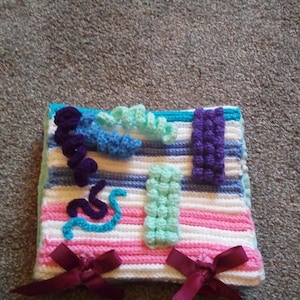May include: A crocheted baby blanket with a variety of textures and colors, including purple, blue, green, pink, and white. The blanket has two bows on the bottom.