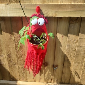 Könnte beinhalten: Ein handgefertigter, roter Häkel-Hängepflanztopf in Form einer Kreatur mit großen weißen Augen und einem roten Kamm. Der Topf enthält kleine grüne Tomatenpflanzen und verleiht jedem Garten oder Außenbereich eine spielerische Note.