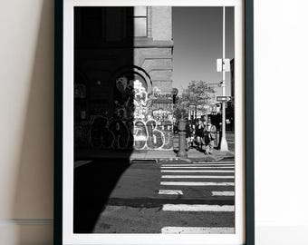 May include: A black and white framed photograph of a city street corner featuring a building with graffiti, a crosswalk, and street signs labeled Broome St. The composition uses high contrast light and shadow to highlight the urban architecture.