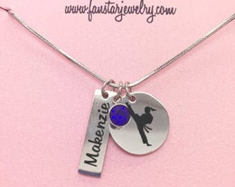 May include: A pink gift box containing a silver necklace. The necklace features a silver charm with a karate silhouette and a blue gemstone. The charm is attached to a silver tag engraved with the word "Makenzie". The box has the text "Fan Star" and "www.fanstarjewelry.com" printed on it. The text "Add your message here" is printed on the bottom of the box.