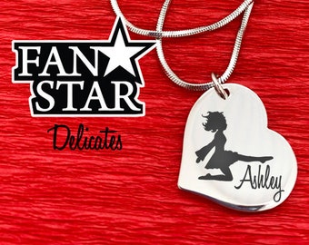 May include: A silver heart-shaped pendant necklace with a laser-engraved silhouette of a dancer and the name "Ashley". The necklace is on a silver chain and comes in a black box with the words "Fan Star" and "Delicates" printed on it.