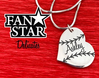 May include: A silver heart pendant necklace with the name "Ashley" and baseball stitching. The necklace is displayed on a red textured surface. The words "Laser Engraved Stainless Steel" are also visible. The necklace's chain is approximately 45 cm long.