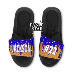 May include: Pair of black slide sandals with blue and waffle-patterned straps. The straps feature the name "JACKSON" and the number "22" with blue icing and red and white sprinkles. The sandals are on a white background.