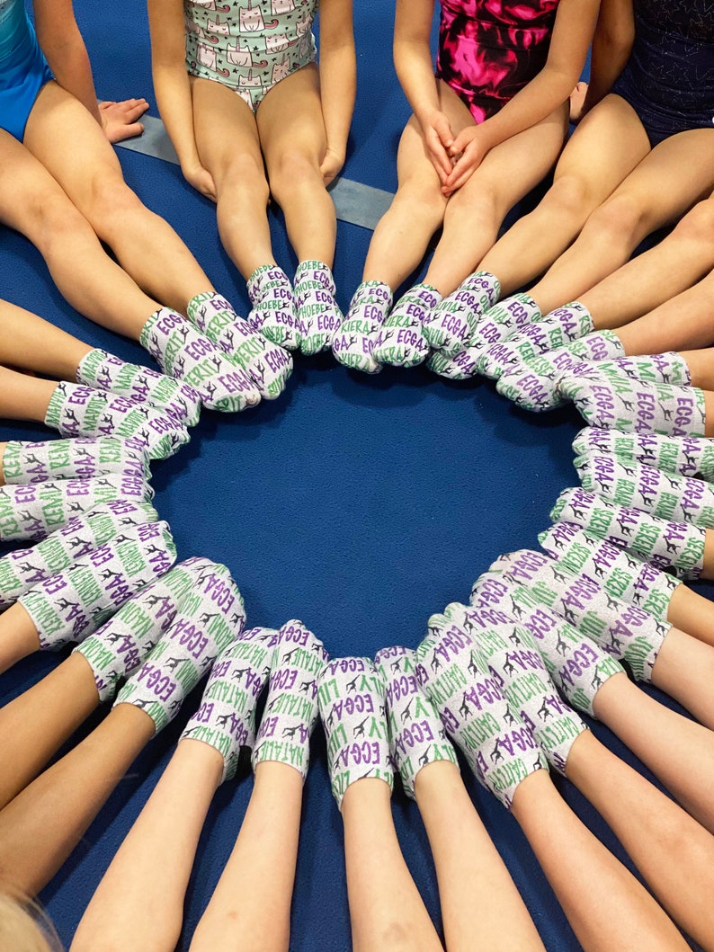 May include: A circle of feet wearing white ankle socks with a repeating pattern of purple and green text and gymnastics silhouettes. The socks feature various names printed in a bold font, including Kiera, Phoebe, Naomi, Natalie, Lily, and Caitlyn.