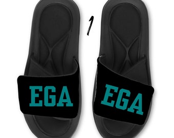 May include: A pair of black slide sandals featuring a wide strap across the top. Each strap displays the text "EGA" in a bold, teal-colored block font. The footbed of the sandals has a contoured design with a textured surface pattern.