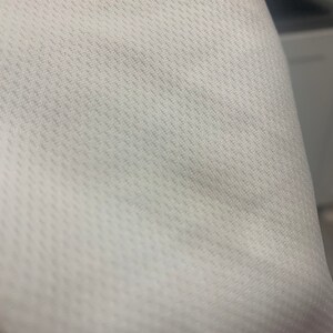 May include: Close-up of a white fabric with a textured pattern. The fabric has a subtle diagonal weave.