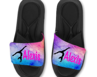 May include: Pair of black slide sandals with a colorful strap featuring a gymnast silhouette and the name "Alexis" in pink. The strap has a watercolor design with pink, blue, and purple hues.