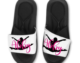 May include: Black and white gymnastic silhouette with the name Abby in pink script on white slides with black straps.