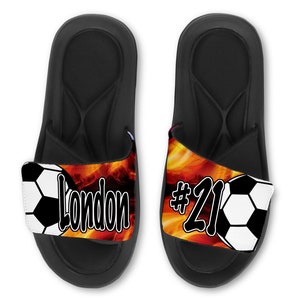 May include: Black slide sandals with a soccer ball and flames design. The left sandal has the name "London" and the right sandal has the number "#21".