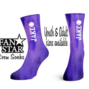 Op de afbeelding: Paarse sokken met een wit baseball en de naam "JAKE" erop gedrukt. De sokken hebben een geometrisch patroon en zijn verkrijgbaar in kinder- en volwassen maten. De sokken zijn gemerkt met het "FAN STAR" logo.