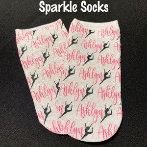 May include: A pair of white glittery ankle socks featuring a repeating pattern of the name Ashlyn in pink script and black silhouettes of a dancer in a leap pose. The text Sparkle Socks is printed in white above the socks.