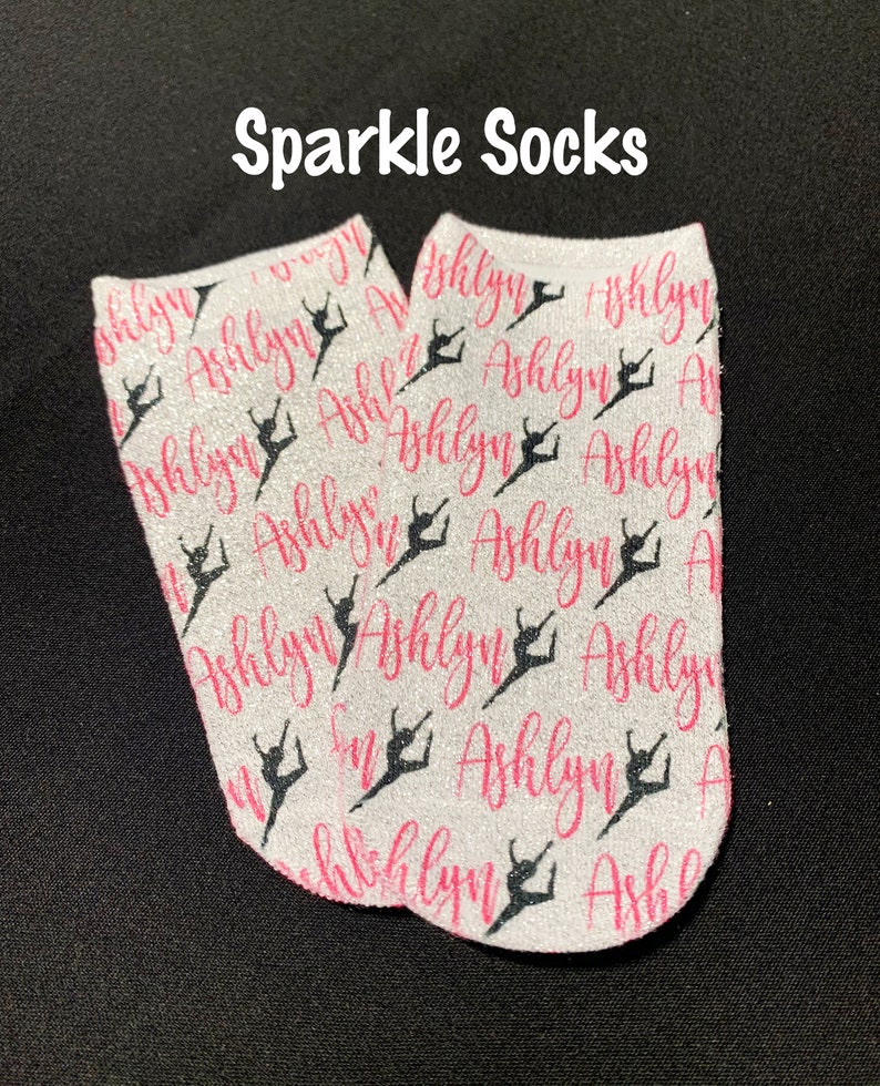May include: A pair of white glittery ankle socks featuring a repeating pattern of the name Ashlyn in pink script and black silhouettes of a dancer in a leap pose. The text Sparkle Socks is printed in white above the socks.