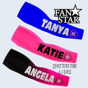 May include: Three arm sleeves in blue, pink, and black, each with a name in white. The blue sleeve says "TANYA", the pink says "KATIE", and the black says "ANGELA". Each sleeve has a heart with a baseball and the number 15. The text "FAN STAR" is at the top.