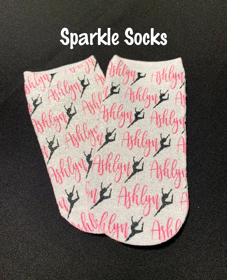 May include: A pair of white ankle socks with a pink glitter finish. The socks feature a repeated pattern of a dancer silhouette and the name "Ashlyn" in pink.