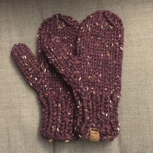 May include: A pair of knitted mittens in a deep burgundy colour with flecks of white and brown throughout. The mittens have a tag that reads "Just for You".