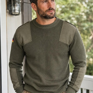 May include: An olive green crew neck sweater featuring ribbed knit fabric, reinforced shoulder patches, and elbow patches. A small German flag emblem is displayed on the right sleeve. The sweater has a classic fit with ribbed cuffs and a ribbed hem.