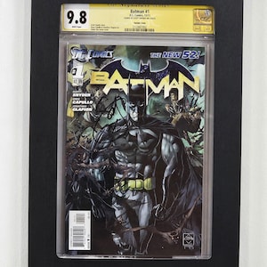 May include: A framed Batman #1 comic book, graded 9.8 by CGC, featuring Batman in a dark, action-packed scene. The cover displays the title "Batman" with a bat symbol, and the text "The New 52!" is also visible.