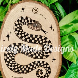 May include: A wood slice with a black and white checkered snake design, stars, and swirls. The snake is curled up with its head facing the right side of the image. The background is a light wood color.
