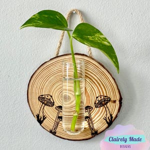 Op de afbeelding: Een houten wanddecoratie met een glazen reageerbuisvaas en een groene plant. Het hout is versierd met een ontwerp van paddenstoelen. De tekst "Clairely Made Designs" is zichtbaar in de rechterbenedenhoek van de afbeelding.