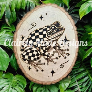 Può includere: Una fetta di legno con un disegno di una rana a scacchi bianchi e neri bruciata a fuoco, con spirali e stelle. La fetta di legno è di colore marrone chiaro.