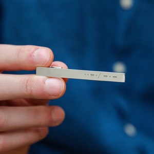 May include: A silver tie bar with a minimalist design featuring a series of dashes and a slash.