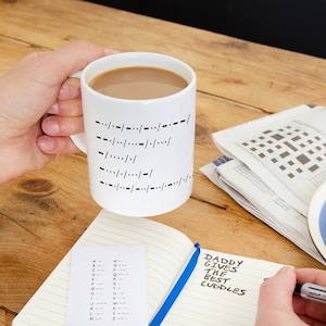 May include: A white mug with black lines on the side, a notebook with the text "DADDY GIVES THE BEST CUDDLES" written in it, and a crossword puzzle in the background.