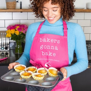 May include: A person wearing a pink apron with the text "bakers gonna bake (bake, bake, bake, bake)" is holding a baking tray with freshly baked cupcakes.