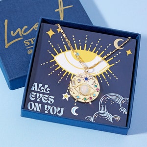 May include: A gold necklace with a large eye pendant featuring colorful gemstones and a white stone in the center. The necklace is displayed in a blue box with the text "All eyes on you" and a crescent moon.