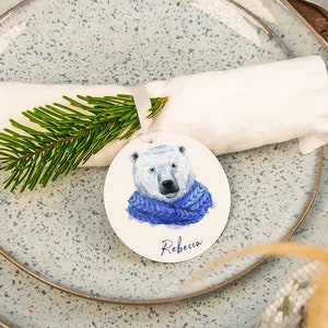 May include: A white napkin with a green pine sprig and a round white place card with a polar bear illustration and the name "Rebecca" printed on it. The place card is on a blue and white speckled plate.