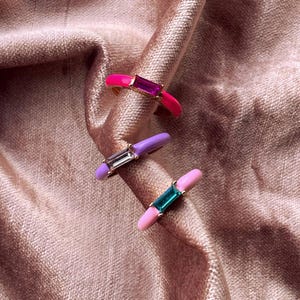 May include: Three colorful rings with rectangular gemstones. The rings feature pink, purple, and light pink enamel bands. Each ring has a different gemstone color: magenta, clear, and teal. The rings are displayed on a pink, textured fabric.