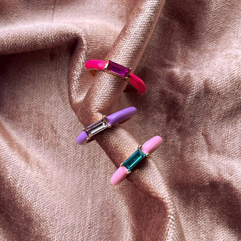 May include: Three colorful rings with rectangular gemstones. The rings feature pink, purple, and light pink enamel bands. Each ring has a different gemstone color: magenta, clear, and teal. The rings are displayed on a pink, textured fabric.