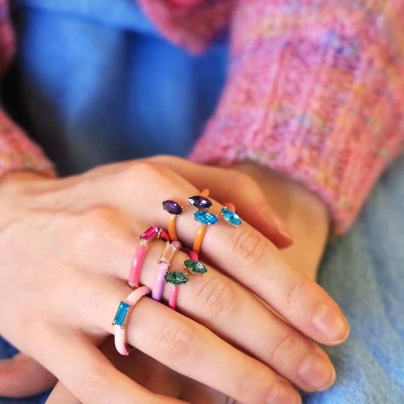 May include: A collection of colorful enamel rings with various gemstone accents. The rings feature a range of colors, including pink, orange, purple, green, and blue. Each ring has a unique gemstone shape and color, adding to the playful design.