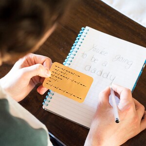 May include: A person is writing "You're going to be a daddy" in a notebook while holding a card with braille dots.