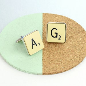 May include: A pair of cufflinks featuring a cream-colored background with black letter tiles. The tiles read "A1" and "G2".