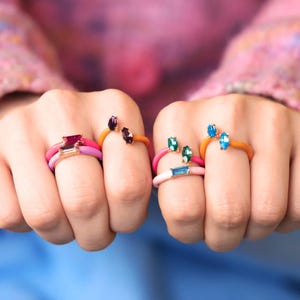 May include: A collection of colorful rings with gemstone accents. The rings feature a variety of colors, including pink, orange, and blue, with gemstones in shades of purple, green, and blue. The rings are displayed on fingers, showcasing their design and vibrant hues.