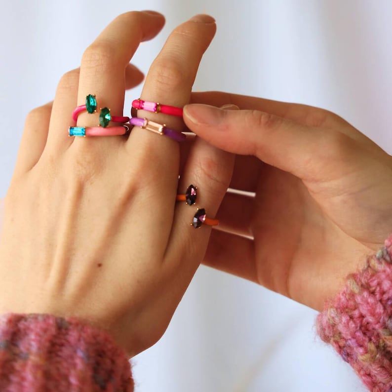 May include: A collection of colorful rings with gemstone accents. The rings feature a variety of colors, including pink, purple, orange, and teal, with rectangular and oval-shaped gemstones. The rings are worn on fingers, showcasing their vibrant design.
