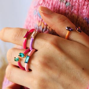 May include: A selection of vibrant enamel rings, each adorned with gemstones. The rings are in shades of pink, purple, and orange, with emerald, amethyst, and clear stone embellishments. The jewellery is displayed on a hand, highlighting the diverse designs.
