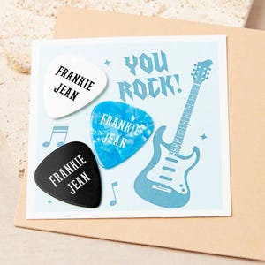 以下が含まれることがあります： 「YOU ROCK！」の文字とギターのイラストが描かれた水色のカード。白、青、黒の3つのギターピックがあり、それぞれに「FRANKIE JEAN」という名前が印刷されています。音符も含まれています。