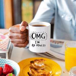 May include: White coffee mug with black text that reads "OMG I'm an adult!" The mug is filled with coffee and being held by a person's hand.