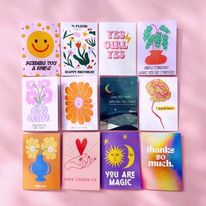 May include: A collection of colorful greeting cards with various designs and messages. The cards feature illustrations of a smiling face, flowers, a plant, a brain, a hand holding a heart, and a sun and moon. Text includes "Sending You A Smile", "Happy Birthday", and "Thanks So Much."