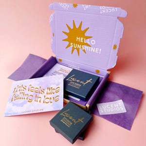 May include: An open, light purple box with the words "HELLO SUNSHINE!" and a sun graphic. Inside are two dark blue jewelry boxes with gold lettering, a card that says "this feels like falling in love", and other purple packaging. The brand name "LUCENT STUDIOS" is visible.