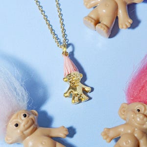 以下が含まれることがあります： ピンクの帽子をかぶった金色のトロール人形ペンダントネックレス、金のチェーン付き。ペンダントは、ピンクと白の髪の毛を持ついくつかのプラスチック製のトロール人形に囲まれており、水色の背景にあります。