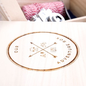May include: A wooden box with a circular design burned into the lid. The design features a compass with arrows pointing in four directions, a bicycle, a mountain, a wave, and a fish. The text "OUR ADVENTURE BOX" is written around the design.