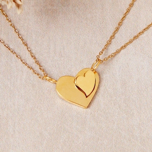 May include: A gold-toned necklace featuring two layered heart-shaped pendants. The larger heart is positioned behind a smaller, slightly offset heart. Both pendants are attached to a delicate gold-toned chain that extends from both sides.
