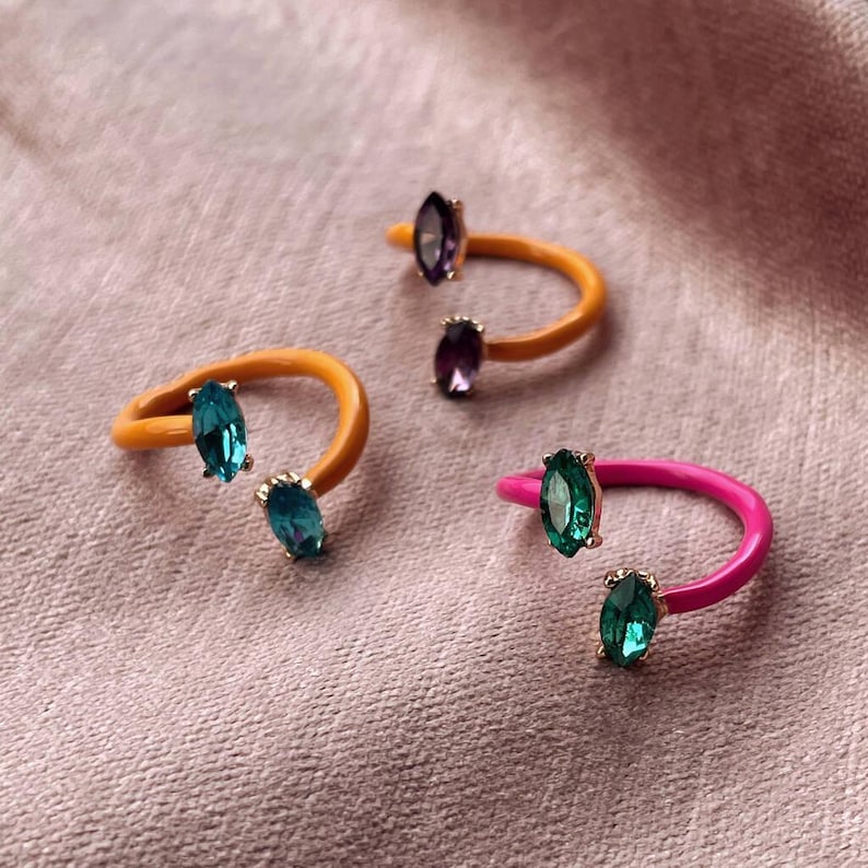 May include: Three colorful rings with gemstone accents. One ring is orange with teal stones, another is pink with green stones, and the third is orange with purple stones. The rings are set against a pink background.