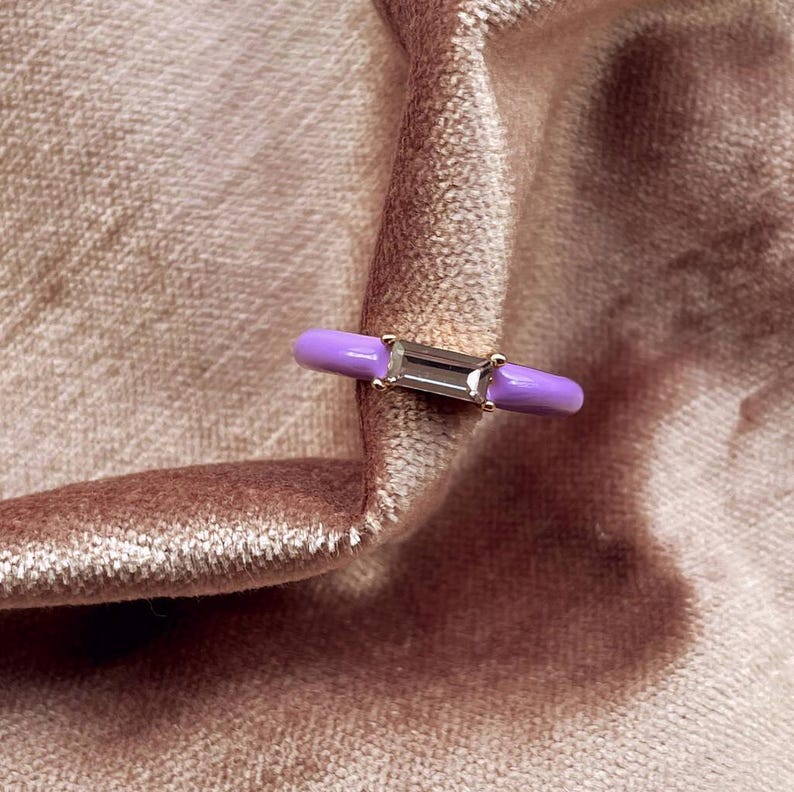 May include: A close-up of a ring with a rectangular, clear gemstone set in a gold band. The ring has a light purple enamel band. The ring is set against a soft, pinkish-brown velvet fabric.